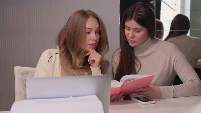 Two females engaged in studying. Women collaborating at desk with books and laptop in office. Two women focused on reading and notetaking in contemporary work environment