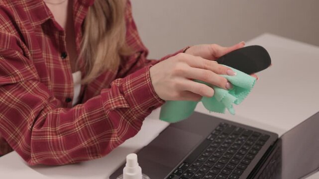 Person wipes black mouse using disinfectant spray and cloth. Closeup of lady sanitizing computer mouse with microfiber cloth and disinfectant spray near keyboard