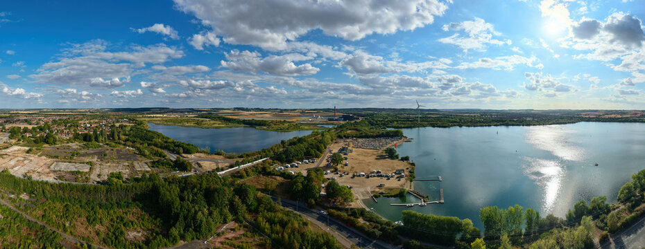 Aerial view of Stewartby Lake featuring calm blue waters, surrounding greenery, a parking area, and distant industrial structures under a bright sky in Bedford, England, United Kingdom.