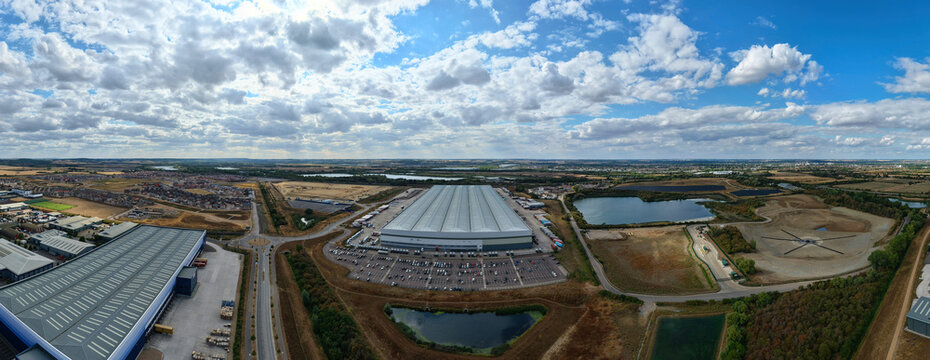 Aerial view of a large logistics hub with industrial warehouses, parked HGVs, and surrounding ponds under a cloudy blue sky in Bedford, England, United Kingdom.