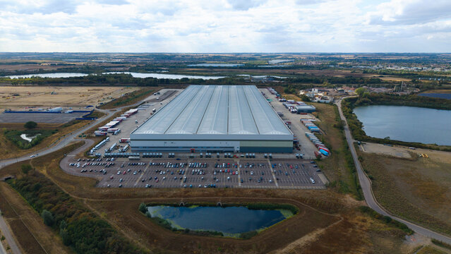 Aerial view of a large logistics hub with industrial warehouses, rows of parked HGVs, and a busy car park, set within a landscape of lakes and fields in Bedford, England, United Kingdom.