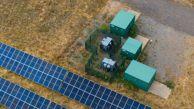 Aerial view of a solar farm featuring rows of photovoltaic panels alongside green battery storage units and electrical transformers within fenced enclosures in Bedford, England, United Kingdom.