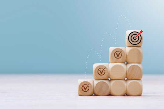 Wooden blocks stacked as steps with checkmarks leading to top goal target icon. Business growth, career ladder progress, milestone achievement, step by step development and strategy concept.