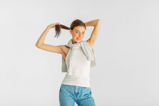 Cheerful model lifts ponytail of her long hair looking into camera, playful energy brands ask for, frame for social media post. Wide shot. Concept of playful mood during studio shooting