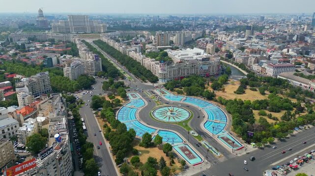Aerial panoramic drone shot of the Unirii Square fountains in Bucharest, Romania, showing urban traffic, green parks, and the Palace of the Parliament in the background on a sunny day.