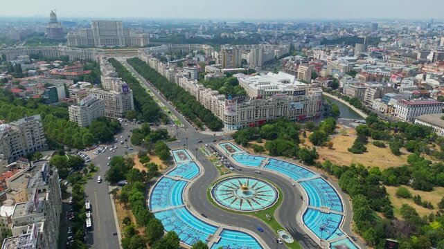 Aerial panoramic drone shot of the Unirii Square fountains in Bucharest, Romania, showing urban traffic, green parks, and the Palace of the Parliament in the background on a sunny day.