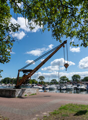 Historic manually operated harbor crane with long metal boom, exposed gear mechanism, chain and lifting hook stands beside a marina filled with boats. Industrial maritime equipment contrasts with mode © Volha
