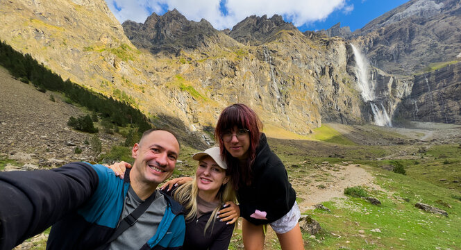 Three people taking a selfie smiling and posing for the camera, in a mountainous landscape with a waterfall in the, with mountains, waterfall, rocky terrain, greenery.