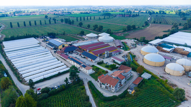 Aerial view of an Italian cattle farm featuring large greenhouses, agricultural buildings, and biogas storage tanks surrounded by lush green fields in Milan, Lombardy, Italy.