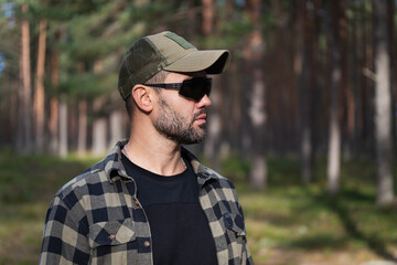 Side profile portrait of a bearded man wearing tactical sunglasses and green mesh cap posing in a pine forest setting. © Dmitri