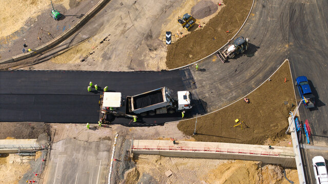 Aerial view of Black Cat Roundabout construction with workers paving fresh asphalt using heavy machinery and a dump truck in Chawston, England, United Kingdom.