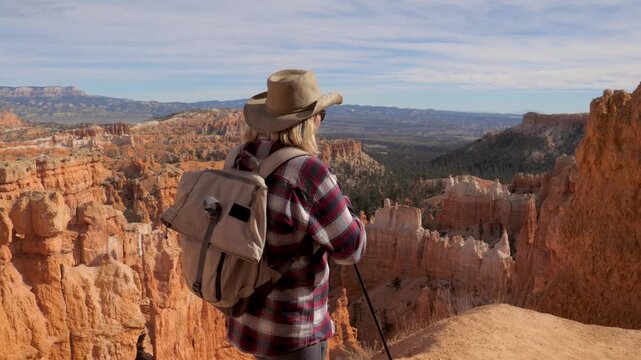 Female explorer with backpack and trekking poles walking on cliff edge, looking out over scenic red rock hoodoos and vast landscape of bryce canyon national park in utah. Following hiker, side view