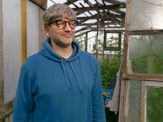 Man stands in wooden greenhouse with glass panels. He wears blue hoodie and glasses and he is in his 40s or 50s with grey beard and hair. Hands on hips, looks at camera. Growing organic produce. © mark_gusev