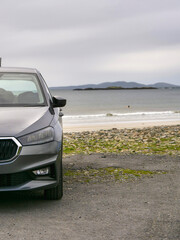 Gray car parked near beach. Ocean waves crash on rocky shore. Distant hills under cloudy sky. Quiet coastal scene with no people. Ideal for travel, adventure, or automotive themes © mark_gusev