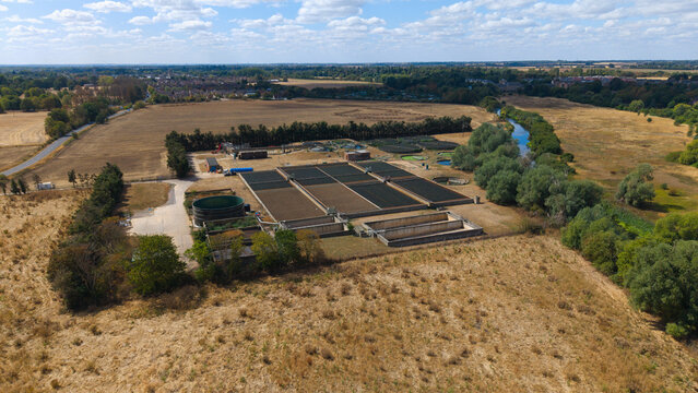 Aerial view of the wastewater treatment works facility featuring settling tanks, filtration beds, and surrounding rural landscape with a river and trees in St Neots, England, United Kingdom.