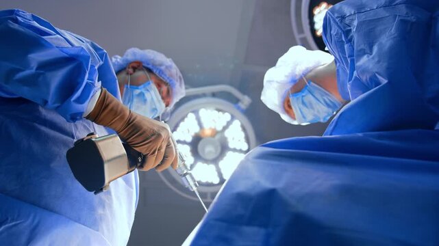 Surgeons handling an instrument from a low viewpoint. Gloved hands hold a metal instrument above blue drapes with a circular lamp in the background.