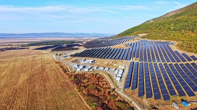A large solar farm spreads over a hillside near a valley with numerous solar panels aligned in rows. The sun shines down, highlighting the layout of the farm and surrounding land.