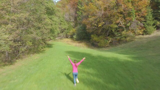 AERIAL: Cheerful young woman runs across a lush green meadow with arms raised toward a colorful autumn forest. Joyful outdoor moment of freedom and connection with nature during a walk in fall season.