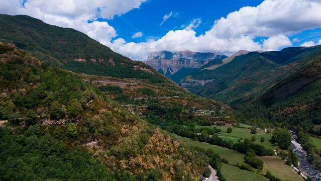 A winding hiking path traverses a vibrant, lush green mountain valley, framed by towering grey peaks and dense forest, under a bright blue sky 