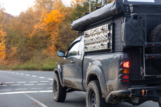 Rugged pickup with expedition camper waits on wet autumn road beside glowing forest, suggesting outdoor escape. Muddy vehicle, travel gear and fall colors evoke adventure, travel and freedom