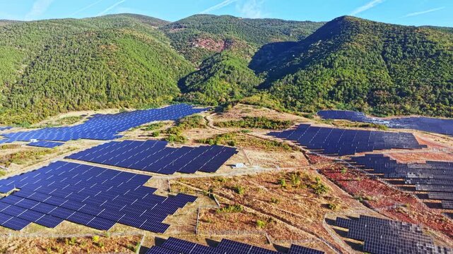 Solar panels cover a vast area in a mountainous region. The panels gather sunlight to generate energy. The scene shows clear skies and green hills in the background.