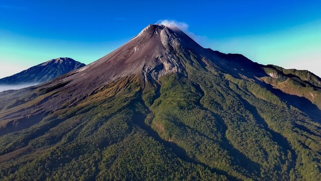 Aerial view of Gunung Merapi showing its volcanic peak, rugged slopes, and surrounding lush green forests in Kecamatan Pakem, Daerah Istimewa Yogya, Indonesia.