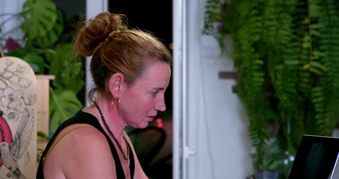 Focused Concentration: A woman with her hair in a bun, intensely focused on a laptop screen, surrounded by lush green plants, suggesting a calm and productive environment.