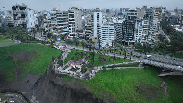 Aerial view of Parque del Amor with its iconic sculpture, lush green gardens, and surrounding modern high-rise buildings along the coastal cliffs in Miraflores, Provincia de Lima, Peru.