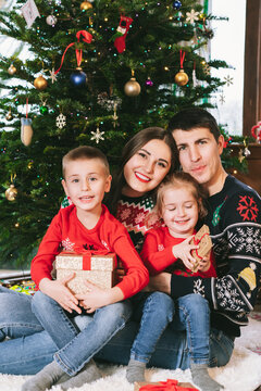 Smiling family portrait in festive sweaters sitting with gifts under Christmas tree at home during holiday. Happy parents and kids cuddling, feeling excited. Family time together. Happy childhood