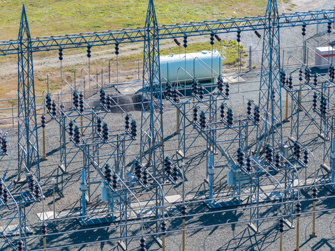 Aerial view of an electrical substation featuring metal lattice towers, high-voltage insulators, and a large storage tank on a gravel ground in Bandon, Oregon, United States.