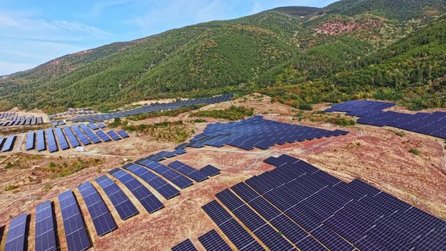 Large solar panels are set up in rows on the ground in a valley. The area is bordered by mountains, and it is sunny. The landscape shows green trees and land.