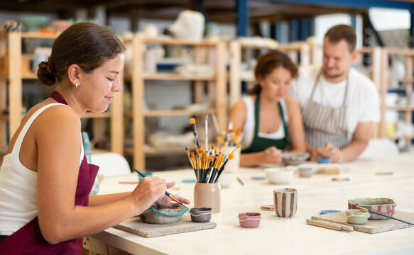 Concentrated girl in apron designing handmade clay plate with brush in taller room