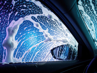 Fototapeta premium Close up view from inside car during automatic wash. White soap bubbles, foam cover windows with blue light. Cleaning process at auto carwash station uses detergent to wash dirty vehicle surface in