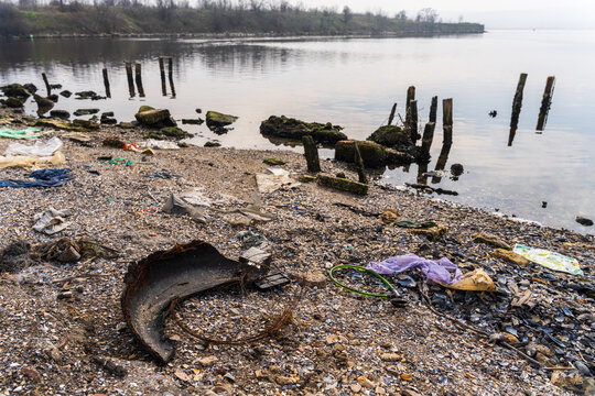 View of the polluted shoreline of Lake Varna with plastic waste, discarded tire, and wooden posts protruding from the water in Varna, Bulgaria.