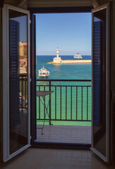 Obraz premium View through open balcony doors toward Chania Lighthouse, turquoise sea and boats in the Old Venetian Harbor of Chania, Crete, Greece.