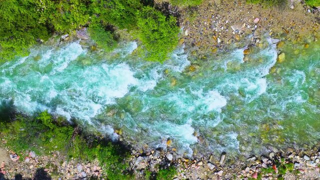Aerial view directly above a clear turquoise mountain stream rushing over rocks with lush green foliage along the riverbanks.