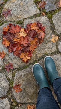 High angle view of person wearing green rain boots standing on a wet stone path next to a heart shape made of colorful autumn maple leaves.