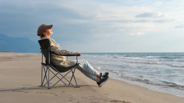 Woman relaxing on beach chair enjoying ocean view. Senior woman wearing casual clothing and cap resting peacefully on beach chair, soaking in expansive ocean scenery with sandy coastline stretching