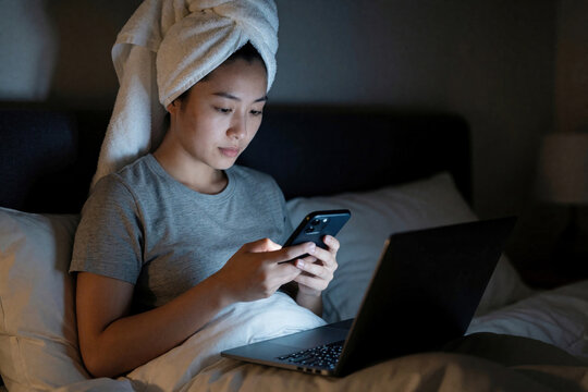Young Asian woman with towel on head sits on bed using smartphone, laptop at night. Female user checks email social media working relaxing in bedroom. Modern digital lifestyle with mobile tech,