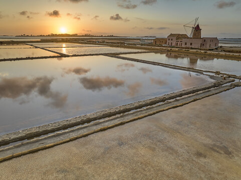 Aerial view of Saline dello Stagnone featuring traditional windmills and salt pans reflecting the golden sunset sky in Marsala, Sicilia, Italy.