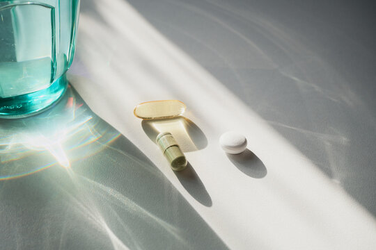 Pills and capsules resting on white surface with glass of water and sunlight