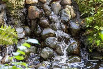Small waterfall cascades over wet stones in forest stream. Fresh mountain water flows through green mossy rocks. Natural river rapid trickles into calm creek pool. © Mati