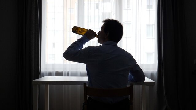 Silhouette of a man sitting alone at a table near a bright window, heavily drinking from a glass bottle, conveying concepts of alcoholism, depression, and social isolation