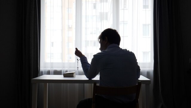 Silhouette of a person sitting at a table near a bright window, holding a rosary and praying with a bible next to them, symbolizing faith and spiritual reflection