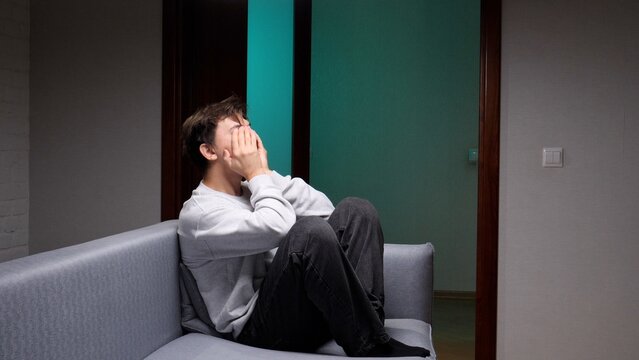 Young man sits alone on a dim sofa, covering his face with his hands, conveying deep sadness, anxiety and isolation in a quiet, somber indoor scene