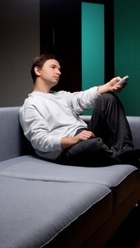 Man relaxing on a comfortable gray sofa in a modern living room, holding a remote control and switching channels, illustrating leisure time, home entertainment, and unwinding after a long day