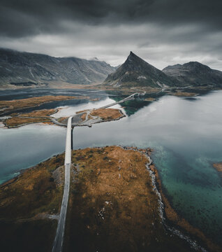 Aerial view of Flakstadbruene bridge connecting islands with rugged terrain, dark water, and dramatic mountains under a cloudy sky in Flakstad, Nordland, Norway.