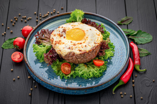 Meat patty topped with a fried egg and lettuce. A yellow yolk sits above the browned meat on a blue plate with leaves and tomato halves.