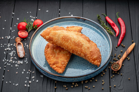 Crisp fried chebureks on plate with spices and peppers. Golden deep cooked turnovers rest on blue ceramic dish surrounded by tomatoes salt and herbs on dark wooden surface.