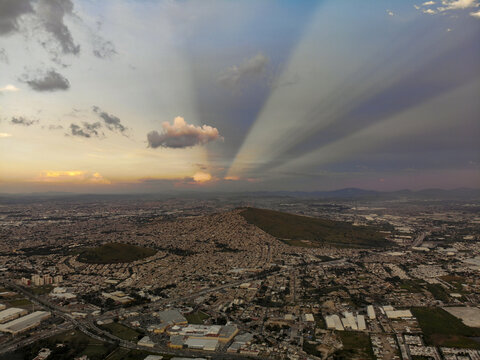 Aerial view of the urban landscape featuring a prominent hill and dramatic crepuscular rays in the sky at sunset in Tlaquepaque, Jalisco, Mexico.